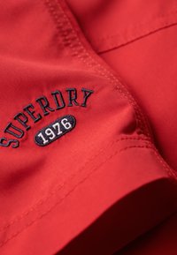 Rotes Stoffdetail mit dunkelblauem, besticktem Text "SUPERDRY" über "1926" in ovaler Form, das die Naht an einem Kleidungsstück zeigt.