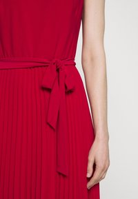 Červené skládání šatů s opásaným páskem. Hladká tkanina, bezrukávový design, viditelné detaily skládání a přiléhavý silueta.