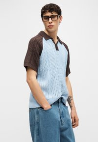 Νεαρό άτομο με σγουρά μαλλιά και γυαλιά, που φοράει μπλε και καφέ πλεκτό κοντομάνικο πουκάμισο και άνετο τζιν παντελόνι με τα χέρια στις τσέπες.