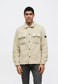 Μπεζ φλις μπουφάν με γιακά, κλείσιμο με κουμπιά μπροστά, δύο τσέπες στο στήθος και μαύρες μεταλλικές λεπτομέρειες. Η υφή φαίνεται μαλακή και ζεστή.