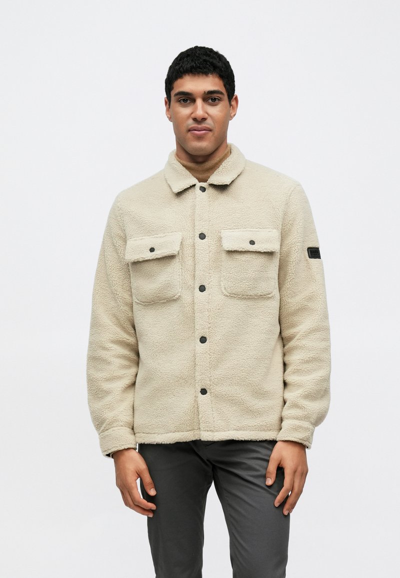 Μπεζ φλις μπουφάν με γιακά, κλείσιμο με κουμπιά μπροστά, δύο τσέπες στο στήθος και μαύρες μεταλλικές λεπτομέρειες. Η υφή φαίνεται μαλακή και ζεστή.