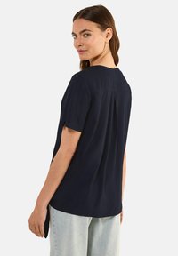 Blusa a maniche corte blu navy con scollo rotondo, dettaglio con piega sul retro e orlo leggermente allungato. Tessuto morbido con una texture liscia.