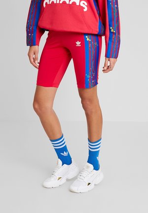Nogi w czerwonych szortach rowerowych Adidas z niebieskimi kwiatowymi paskami, niebieskie skarpetki Adidas, białe buty sportowe Adidas i część czerwonej bluzy Adidas.