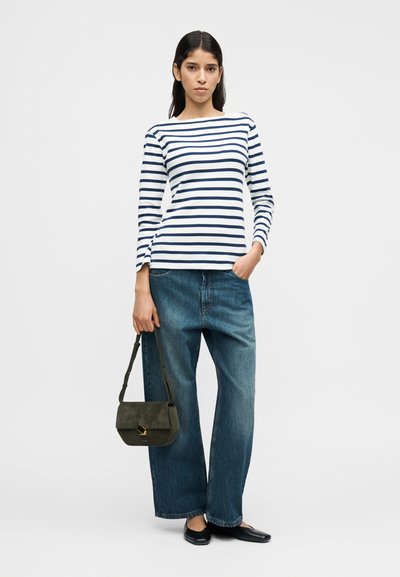 Ριγέ μακρυμάνικη μπλούζα σε λευκό και ναυτικό μπλε, συνδυασμένη με φαρδιά μπλε τζιν παντελόνια. Το μοντέλο κρατά μια τσάντα χειρός από σουέτ σκούρο πράσινο με χρυσά διακοσμητικά.