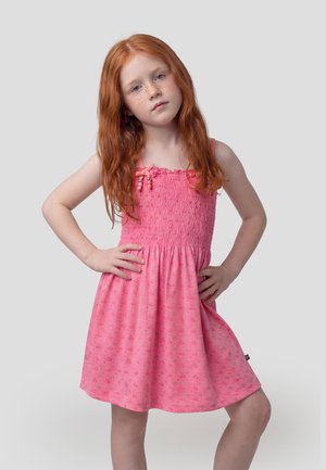 Niña pelirroja con pecas, de pie con las manos en las caderas, vestida con un vestido rosa sin mangas sobre un fondo claro y liso.