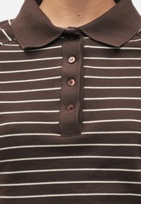 Polo marron avec des rayures blanches horizontales, doté d'un col et de quatre boutons marron sur le devant. Tissu en coton texturé.