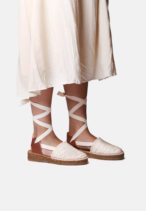 Jalad, mis kannavad beeže espadrille-stiilis sandaale, millel on pahkluul kinnitatavad paelad, ning voolava kreemika midi-seeliku taustaks valge taust.