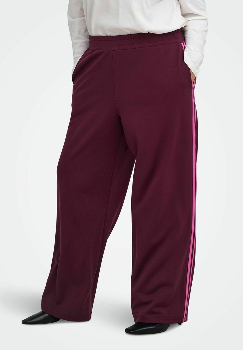 Personne portant un pantalon large violet foncé avec des bandes roses latérales, un haut blanc à manches longues et des chaussures noires pointues, les mains dans les poches.