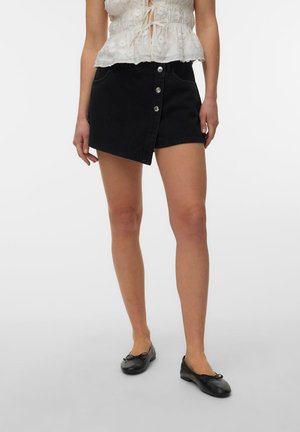 Femme portant une mini-jupe noire asymétrique boutonnée, un top blanc sans manches à lacets et des ballerines noires, debout sur un fond uni.