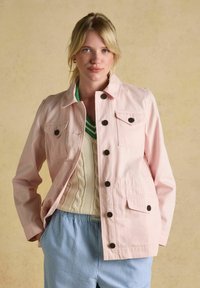 Lyshåret kvinde iført en lyserød jakke med knapper over en cremefarvet kabelstrikket vest og blå stribede bukser, stående mod en beige baggrund.