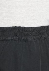 Calças de tecido preto com uma cintura elástica, apresentando uma textura suave e um ajuste descontraído. Termina logo acima da bainha de uma blusa de cor clara.