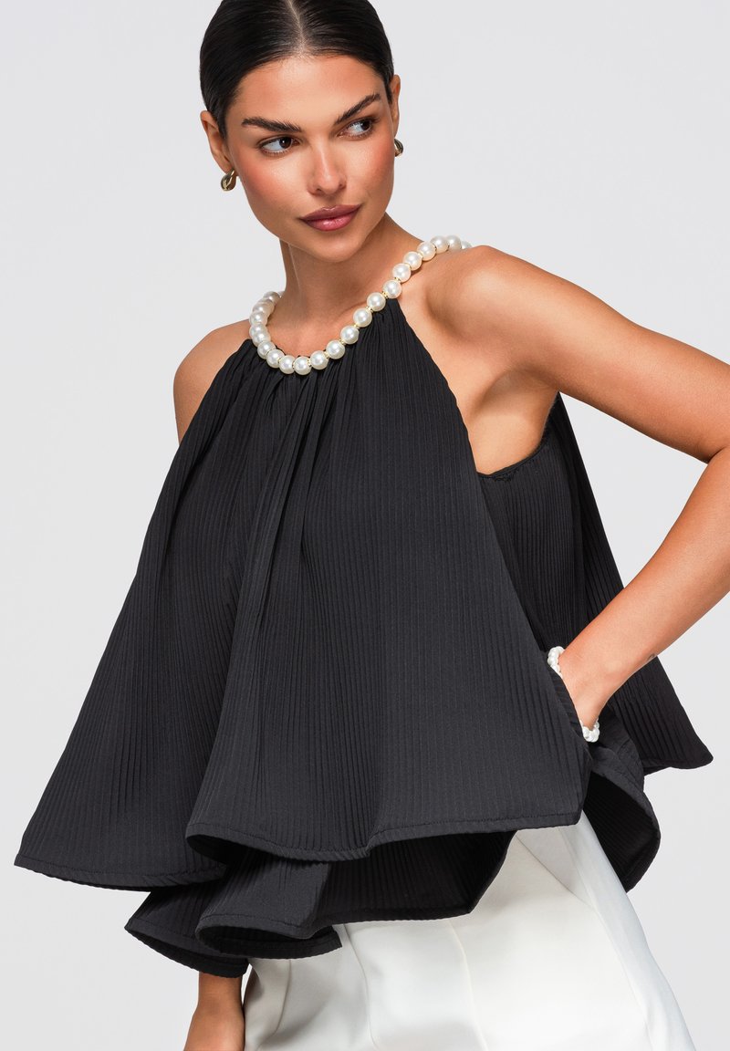 Femme portant un haut dos nu plissé noir ample avec un grand collier de perles et un bracelet assorti, associé à un pantalon blanc, posant la main dans la poche.