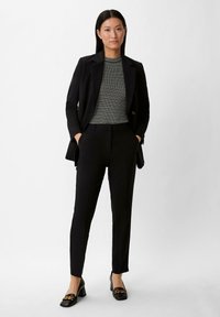 Damen-taillierte schwarze Blazer über einem gestreiften Top, enganliegenden schwarzen Hosen und hohen Loafers mit goldenen Kettenakzenten. Weicher Stoff, elegantes Design.