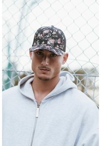 Fekete baseball sapka virágmintával, a logó a elején látható, világosszürke cipzáras pulóverrel viselve, láncsoros háttér előtt.