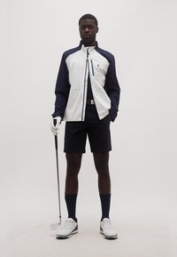 Hombre vestido con una chaqueta de golf blanca y azul marino, pantalones cortos negros, guante de golf blanco, calcetas hasta la rodilla y zapatos de golf blancos, sosteniendo un palo de golf.