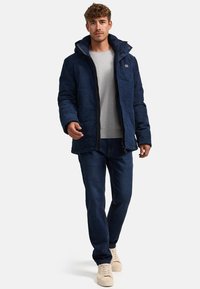 Marineblaue Steppjacke mit Kapuze, Frontreißverschluss und strukturierter Stoffoberfläche, kombiniert mit einem hellgrauen Pullover und dunkelblauen Jeans.