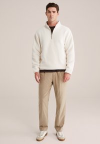 Pullover in pile a collozzo con zip, di colore bianco sporco, caratterizzato da una consistenza morbida, con polsini a costine, indossato sopra una camicia marrone scuro, abbinato a pantaloni beige e sneakers.