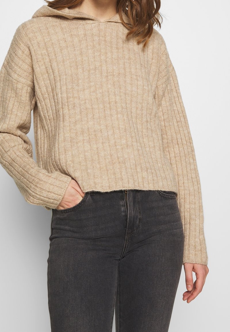 Femme portant un pull beige en maille côtelée avec capuche et manches longues, associé à un jean noir délavé taille haute, main dans la poche.