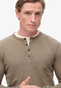 Lys brun langærmet skjorte med henley-design, der har rund hals, seks sorte knapper og et blødt, tekstureret stof.