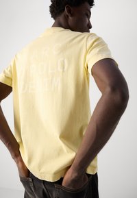 Yellow cotton polo shirt featuring a matte white logo print on the back. Short sleeves and a classic collar. The model has hands in pockets.
