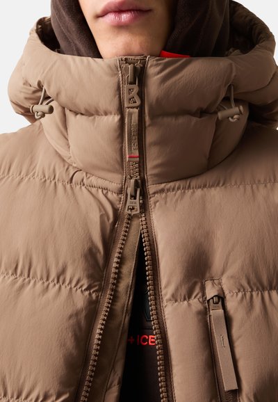 Hellbraune Pufferjacke mit hohem Kragen, ausgestattet mit einem durchgehenden Reißverschluss und verstellbaren Schnüren. Innenlage sichtbar mit roten Akzenten.