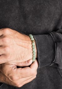 Bracciale con perline di pietra verde con un accento dorato, indossato su un polso, caratterizzato da una superficie liscia e perline rotonde su una felpa scura.