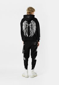 Sudadera negra con estampado de alas plateadas en la parte posterior, combinada con pantalones negros con detalles desgastados y un bolsillo lateral. Botas blancas.