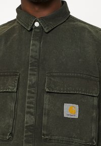 Zielona koszula dżinsowa z kołnierzykiem zapinanym na guziki, dwoma kieszeniami na piersiach oraz naszywką z logo Carhartt. Teksturowana tkanina o nieco wytartym wyglądzie.