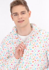 Giacca a vento colorata con sfondo bianco e motivi di foglie multicolori; chiusura a zip frontale e colletto regolabile.