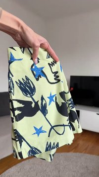 Χέρι που κρατά διπλωμένο κίτρινο ύφασμα με μοτίβα μαύρου πουλιού και μπλε αστέρι, σε σκηνικό καθιστικού.