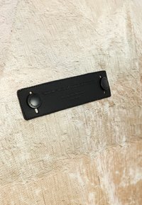 Tejido texturizado en tono marrón, con una etiqueta de cuero que presenta herrajes negros, texto grabado y broches circulares. Patrón prominente en el material.