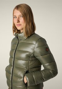 Giacca puffer verde oliva con superficie lucida, chiusura zip, tasche laterali e colletto alto. Presenta un design trapuntato e un logo sulla manica.