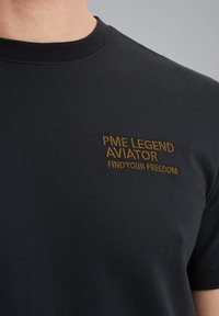 Černé tričko s krátkým rukávem vyrobené z texturované látky. Na hrudi se nachází zlatá výšivka s nápisem "PME LEGEND AVIATOR FIND YOUR FREEDOM".