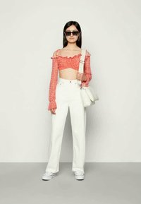 Mulher vestindo um cropped vermelho com ombros à mostra, calças brancas de cintura alta, tênis brancos, óculos de sol pretos e carregando uma bolsa branca de ombro.