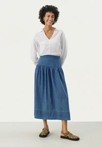 Denim maxi szoknya, illeszkedő derékkal és ráncolt részletekkel, laza, fehér leninggel és barna szandállal párosítva.