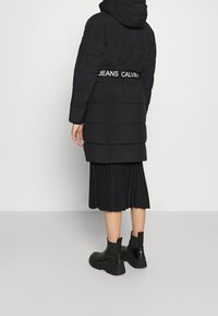 Černý zateplený kabát s kapucí, zdobený bílým logem na pásku. Dole se outfitu přidává černá plisovaná sukně a robustní černé kotníkové boty.