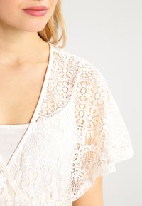 Vit spetsblus med V-ringning, prydd med intrikata blommönster och luftiga ärmar. Texturerad tyg förbättrar den visuella detaljrikedomen.