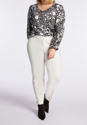 Vrouw draagt een zwart-witte blouse met lange mouwen met patroon, witte nauwsluitende broek en zwarte enkellaarzen met puntige neus, staat met één hand langs het lichaam.