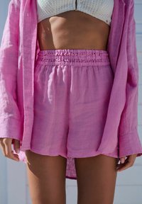 Kvinna i en vit texturerad crop top, lösa rosa högmidjade shorts med draperad midja, och en matchande rosa långärmad skjorta öppen.