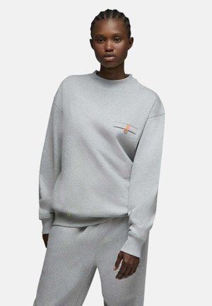 Femme portant un sweat-shirt gris clair et un pantalon de survêtement assorti, debout les mains le long du corps sur un fond blanc uni.