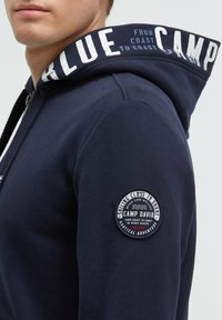 Sudadera con cremallera en color marino y capucha con cordón, que presenta texto blanco en la capucha y un parche circular en la manga izquierda. Material de algodón.