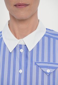 Gros plan d’une personne portant une chemise rayée verticalement bleu et blanc avec un col pointu blanc et un rabat de poche triangulaire boutonné.