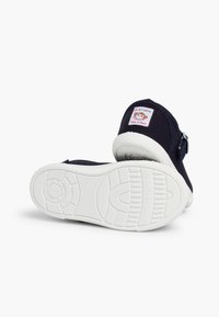 Scarpe di tela blu navy con suola in gomma bianca e una fibbia blu, caratterizzate da un patch con logo animale colorato sul tallone.