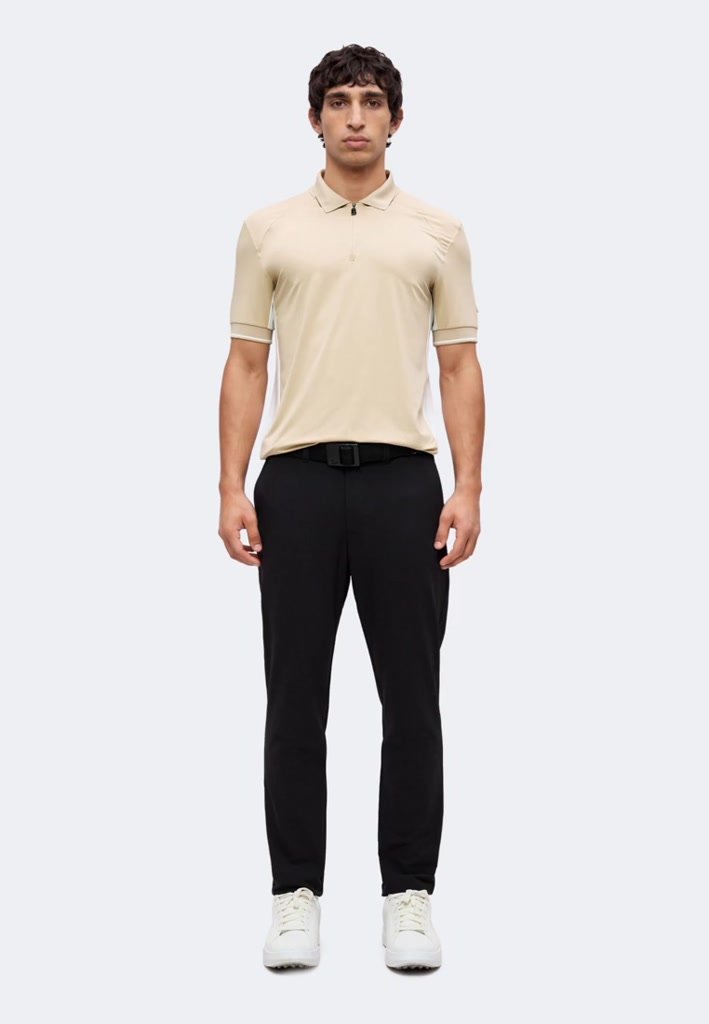 Homme debout, face à l'avant, portant un polo beige à manches courtes, un pantalon noir avec ceinture et des baskets blanches sur un fond uni.