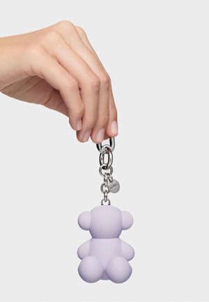 Ręka trzyma srebrny brelok na łańcuszku z małym, lawendowym zawieszonym w kształcie misia, na białym tle.
