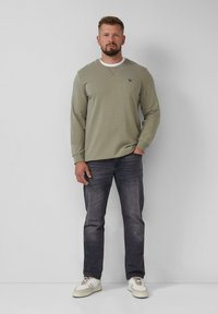 Uomo in piedi contro un muro chiaro, indossando un felpa verde oliva, jeans scuri e sneakers bianche, con una mano in tasca.