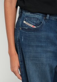 Blugi din denim într-o nuanță albastru închis cu textură ușor uzată. Etichetă cu logo vizibilă în buzunarul din spate, evidențiind detaliile brandului.