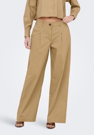 Donna che indossa pantaloni beige a vita alta e gamba larga abbinati a una camicia beige corta con bottoni e tacchi aperti marroni.