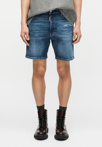 Homme portant un short en jean bleu délavé, des chaussettes foncées et des bottines brillantes en cuir marron foncé à lacets, debout avec les mains détendues le long du corps.
