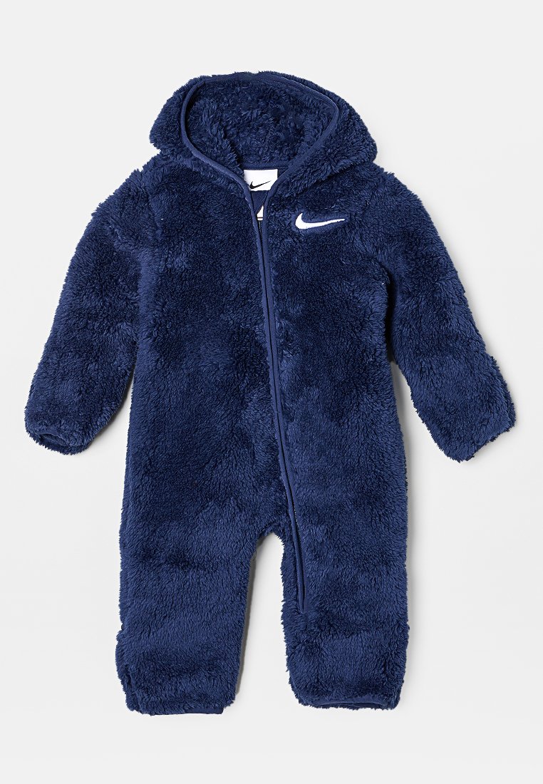 Marinblå fleeceoverall med mjuk textur, full dragkedja och huva. Har en vit Nike-logotyp på bröstet och långa ärmar.